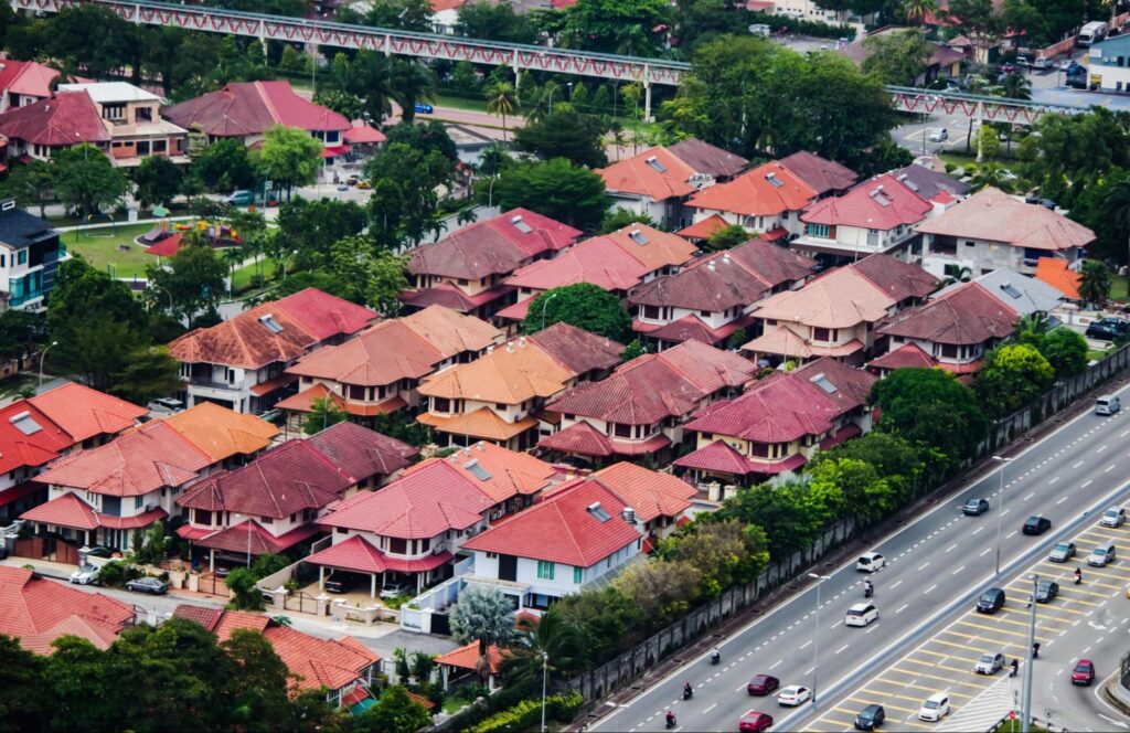 An aerial view shows a residential neighborhood in Malaysia with red-roofed landed houses lined along a busy road