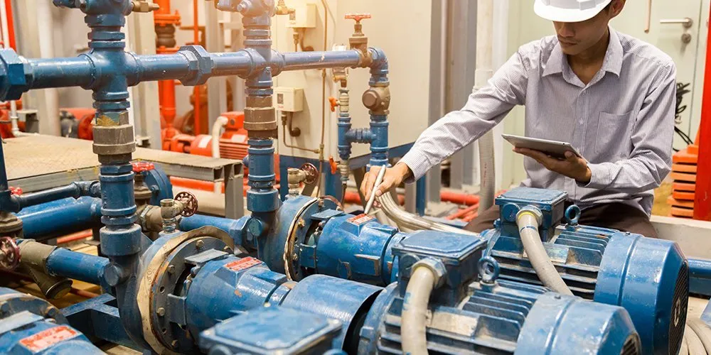 A worker in a hard hat inspects industrial blue pumps and piping while holding a tablet