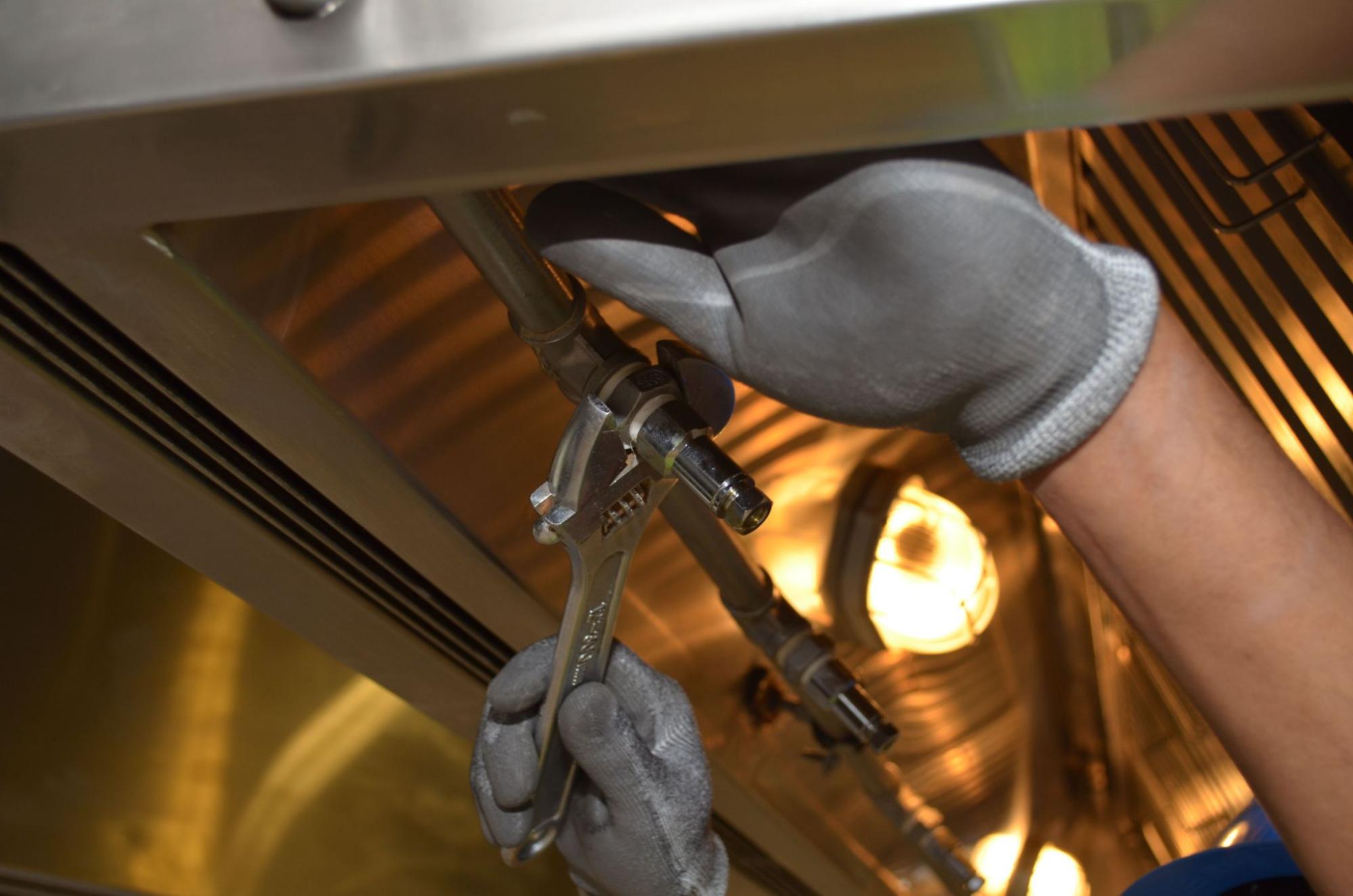 A gloved hand uses a wrench to tighten a pipe fitting under stainless steel equipment