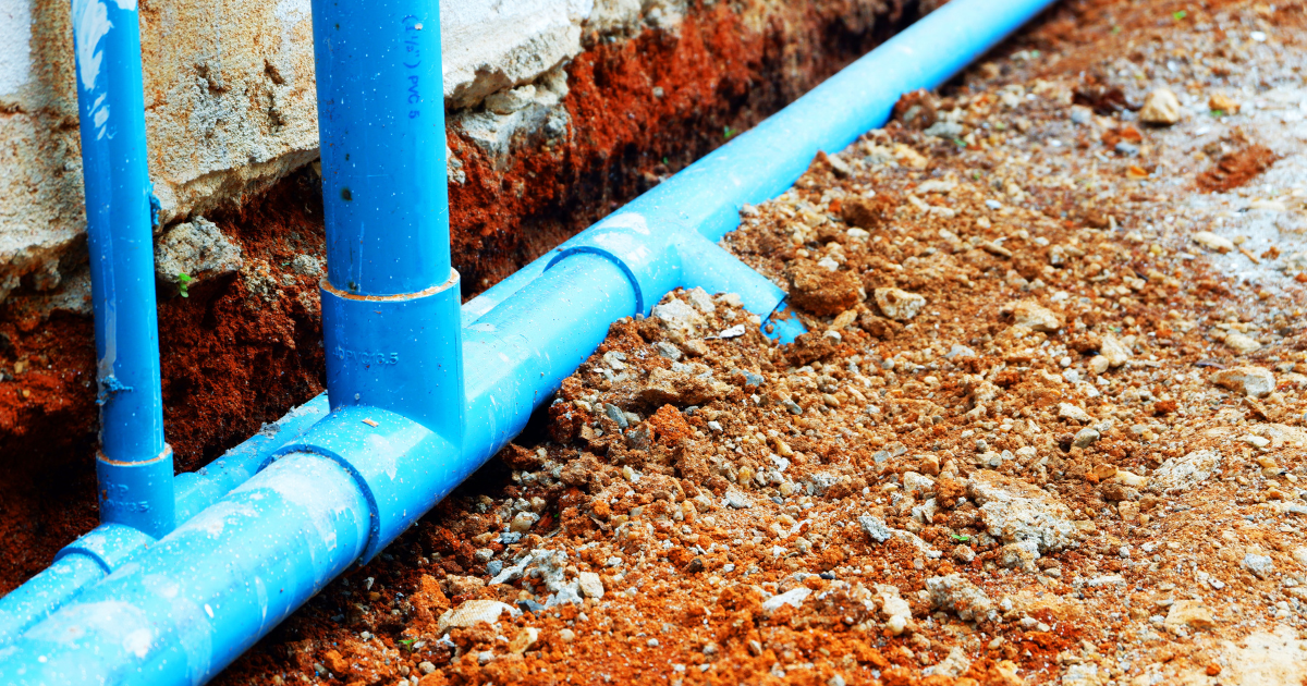 Blue PVC pipes are laid in a trench alongside a building's foundation.